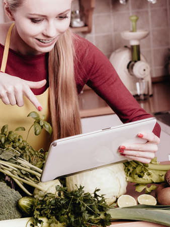 Young woman in kitchen having many green vegetables on table, holding tablet thinking about cooking something and searching for recipes in internetの写真素材
