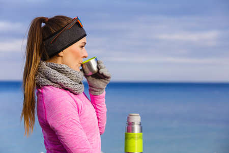 Woman athlete fit girl wearing warm sporty clothes outdoor in cold weather drinking hot tea from vacuum flask thermos, warming up. Sports, activities, hiking in winter or autumn timeの写真素材