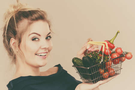 Buying good food, vegetarian products. Positive woman holding shopping basket with green red vegetables inside, recommending healthy high fibre diet, lifestyle modification, on greyの写真素材