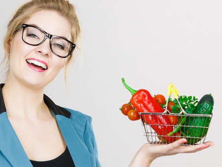 Lose weight, buying healthy food, vegetarian products. Positive business woman in glasses holding little shopping basket with vegetables, on greyの写真素材