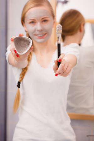 Skin care. Young woman holding in hands brush and clay mud mask to apply on skin face, in bathroom. Girl taking care of her oily compexion. Spa wellbeing.の写真素材