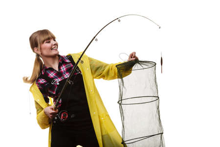 Spinning, angling, cheerful fisherwoman concept. Happy woman in yellow raincoat holding fishing rod and keepnet having fun.の写真素材