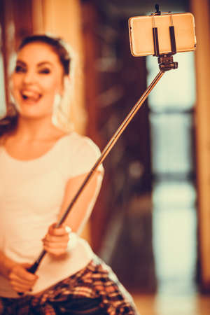 Happy cute pretty woman taking self photo with selfie stick. Attractive young girl with long hair wearing white shirtの写真素材
