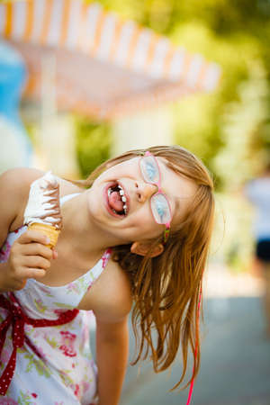 Summertime joy, summer recreation outside concept. Toddler girl in eyeglasses eating ice cream, having fun making funny faces.の写真素材