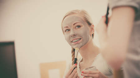 Skincare. Blonde woman in bathroom with gray clay mud mask on her face. Young lady taking care of skin. Spa beauty wellness. Filtered photoの写真素材