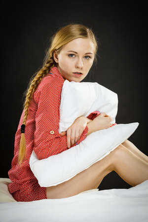 Mental health depression insomia concept. Sad depressive young woman teen blonde girl wearing red pajamas sitting on bed embracing pillow, on dark black night backgroundの写真素材