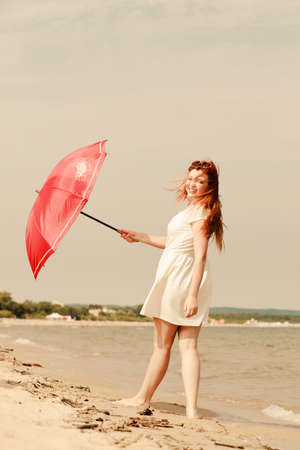 Redhead woman wearing gauzy white dress walking on beach holding red umbrella, having nice relaxing summertime walkの写真素材