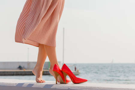 Hobby, idyllic aspects of femininity concept. Woman walking on jetty without shoes wearing beautiful long light pink dress, high heels standing nex to legs.の写真素材