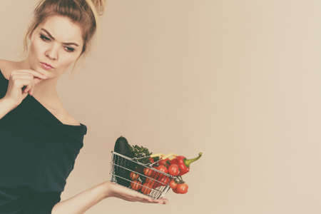 Adult woman with raw food, questioning healthy lifestyle recommendations, origin vegetagles. Female holding small shopping basket with products, thinking face expressionの写真素材