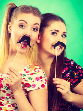 Two happy women holding fake moustache on stick having fun wearing tshirts with flower pattern. Photo and carnival funny accessories concept.の写真素材