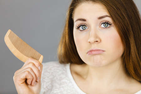 Attractive young woman having brown hair being confused holding comb. Studio shot on grey...