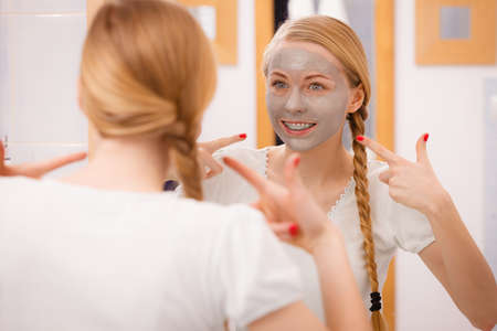Skincare. Blonde woman in bathroom with gray clay mud mask on her face. Young lady taking care of skin. Spa beauty wellness.の写真素材