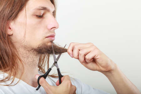 Cut and shave concept. Young man with long beard holding scissors. Boy cutting hair on chin.の写真素材