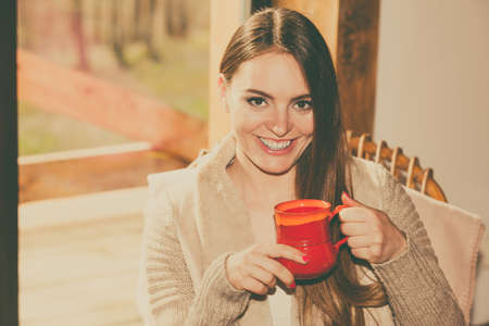 Calm and coziness. Young woman at home sitting comfortable on rocker chair in front of window relaxing in her living room enjoying coffee or tea drinkの写真素材