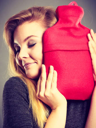 Positive happy woman hugging warm hot water bottle in red soft fleece cover, on grey. Health care, pain relievers, treatment objects concept.の写真素材