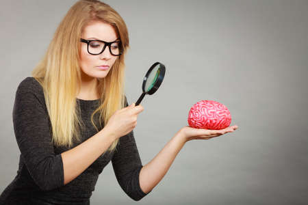 Blonde woman holding magnifying glass investigating something and looking closely at fake brain.