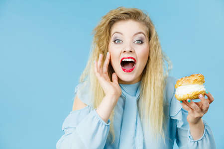 Sweet food and happiness concept. Funny joyful blonde woman holding yummy choux puff cake with whipped cream, excited face expression. On blueの写真素材