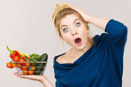 Adult woman do not like to eat raw food, questioning healthy lifestyle recommendations, origin vegetagles. Female holding small shopping basket with products, displeased shocked face expressionの写真素材
