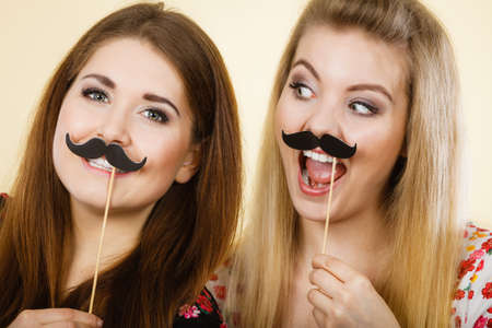 Two happy women holding fake moustache on stick having fun wearing tshirts with flower pattern. Photo and carnival funny accessories concept.の写真素材
