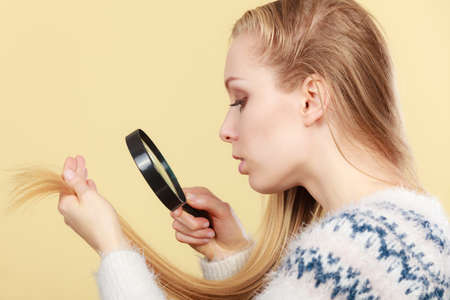 Haircare, health problem concept. Unhappy blonde woman looking at ends of her blonde hair through magnifying glassの写真素材