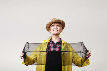 Spinning, angling, cheerful fisherwoman concept. Happy woman in yellow raincoat holding empty fishing keepnet, having fun.の写真素材