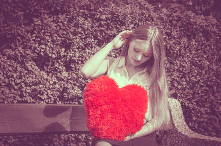 Love and feelings. Unhappy hurt woman sitting on bench with big red heart in hands. Beautiful sad girl in park.の写真素材