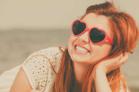 Summer relaxation and recreation concept. Happy redhead adult woman wearing sunglasses in heart shape lying on beach during summertime.の写真素材