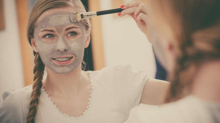 Facial dry skin and body care, complexion treatment at home concept. Happy young woman applying grey mud mask on her face with brush.の写真素材