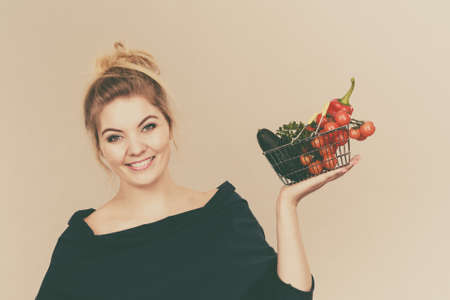 Buying good food, vegetarian products. Positive woman holding shopping basket with green red vegetables inside, recommending healthy high fibre diet, lifestyle modification, on greyの写真素材