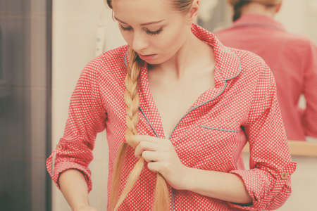 Fancy trendy blond hairstyle at home concept. Woman wearing pink pajamas doing braid on blonde hair