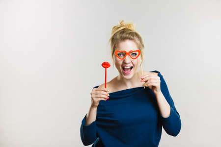 Happy blonde woman holding carnival accessories on stick fake red lips and paper heart shaped...