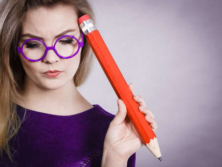 Person confused thinking seeks a solution. Pensive thoughtful student girl or business woman female teacher wearing nerdy glasses coming up with an idea, holding big red pencil. Studio shot on grey.の写真素材