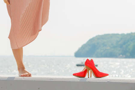 Hobby, idyllic aspects of femininity concept. Woman walking on jetty without shoes wearing beautiful long light pink dress, high heels standing nex to legs.の写真素材