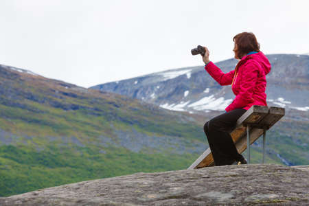 Tourism adventure and travel. Female tourist hiker sitting on bench in stone mountains taking photo with camera, looking at scenic view, Norway Scandinavia.の写真素材