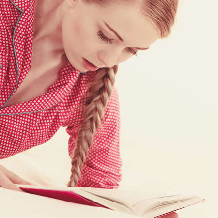 Girl lying in bed reading book. Young blonde female wearing red dotted pajamas relaxing at home on mattress.の写真素材