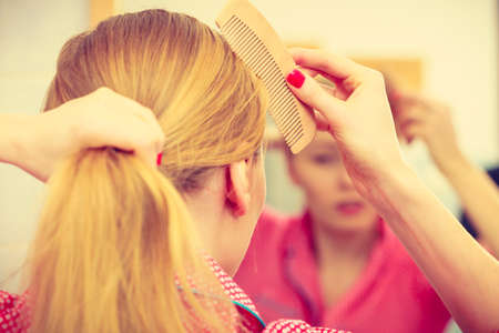 Woman combing brushing her long blonde smooth hair in bathroom, looking in mirror. Teen girl taking care refreshing her hairstyle in morning. Haircare concept.の写真素材