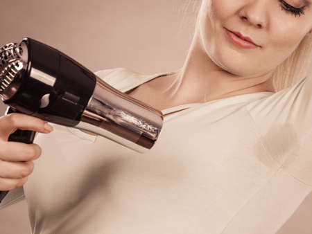 Woman drying wet armpit using hair dryer. Getting rid of sweat and bad smell, hyperhidrosis.のeditorial素材
