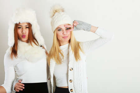 Fashion winter outfit concept. Two girls blonde and mulatto in warm white clothing having fun. Attractive women wearing fur caps, scarfs, gloves.の写真素材