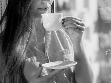 Young woman sitting on windowsill looking through window enjoying her free time, relaxing while drinking coffee or tea from cup.の写真素材