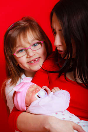 Toddler daughter and mother holding little newborn baby with red background. Family love, childhood and parenthood concept.の写真素材