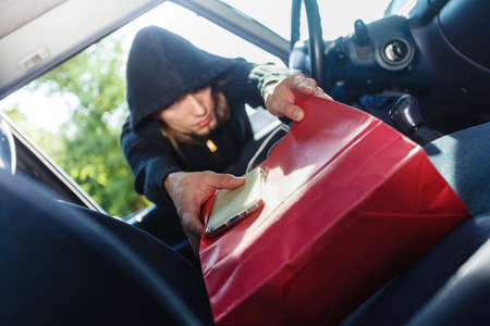 Anti theft system problem concept. Burglar thief man wearing black clothes breaking into car, stealing smartphone and red shopping bagの写真素材