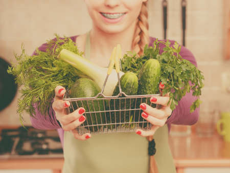 Buying healthy dieting food concept. Woman in kitchen having many green vegetables holding small shopping basket.の写真素材