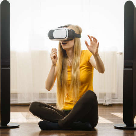 Young woman wearing virtual reality goggles vr 3d box sitting on floor in living room, listening to...