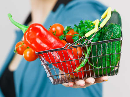 Lose weight, buying healthy food, vegetarian products. Woman hand holding little shopping basket with high fibre red green vegetables inside, on greyの写真素材