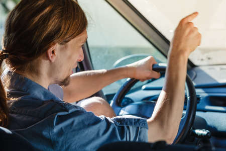 Traffic jam, anger concept. Young furiously angry man wearing jeans shirt having long hair, driving car and screaming at other driversの写真素材