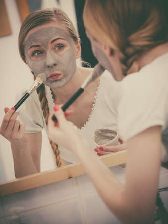 Facial dry skin and body care, complexion treatment at home concept. Young woman applying grey mud mask on her face with brush.の写真素材