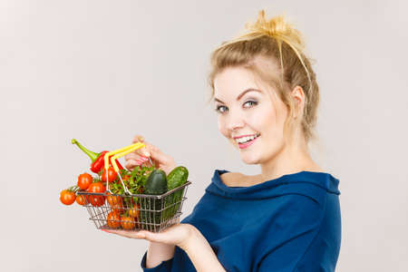 Buying good food, vegetarian products. Positive woman holding shopping basket with green red vegetables inside, recommending healthy high fibre diet, lifestyle modification, on greyの写真素材