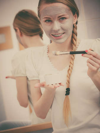 Skincare. Blonde woman in bathroom applying with brush gray clay mud mask to her face. Young lady taking care of skin. Spa beauty wellness.の写真素材