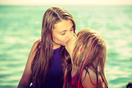 Mother and daughter kissing near sea rock with water in background on sunny dayの写真素材