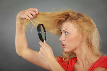 Haircare, health problem concept. Unhappy woman looking at ends of her blonde hair through magnifying glassの写真素材
